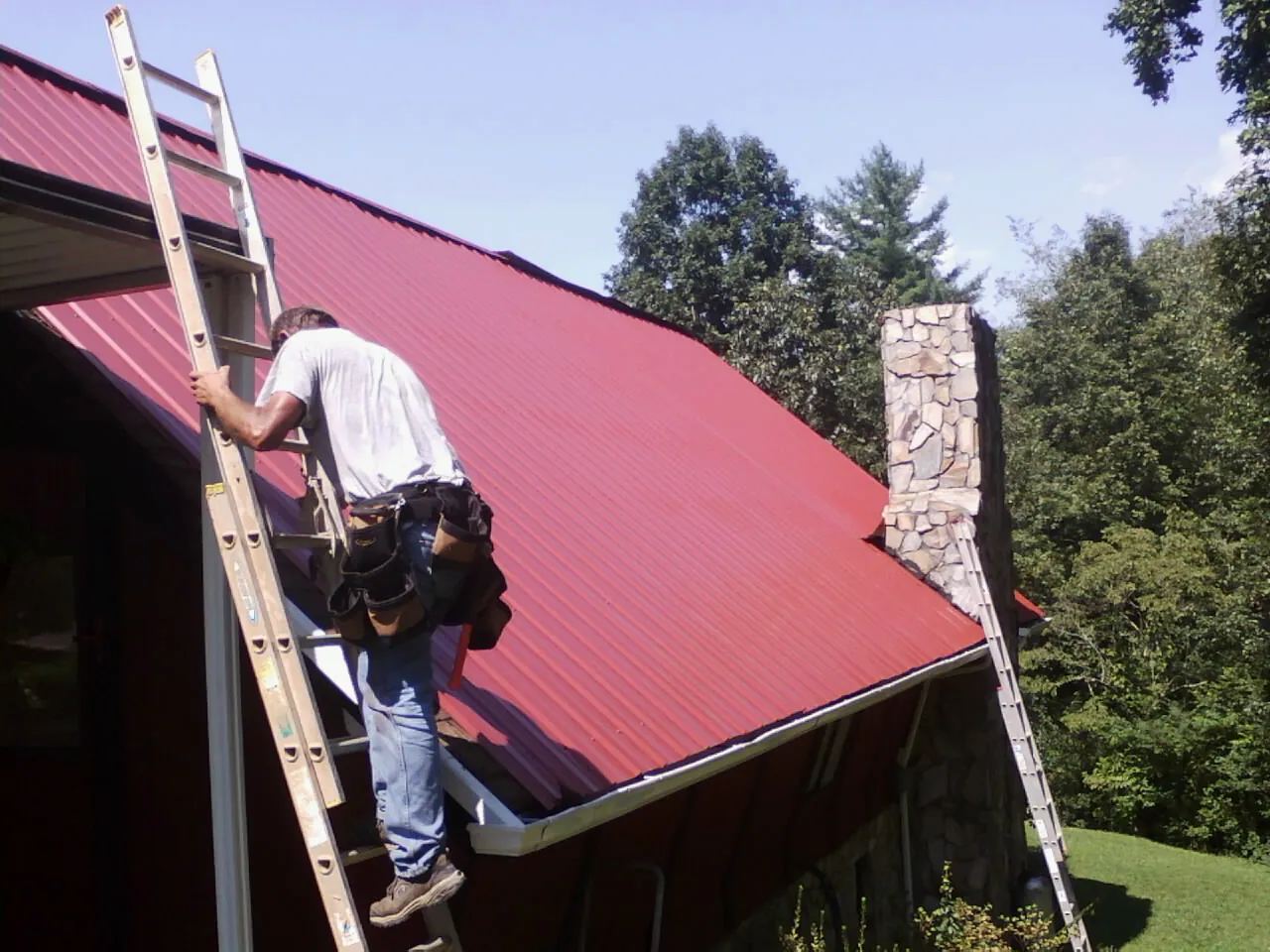 Roofer on a ladder beside a red metal roof and stone chimney — Soffit & Fascia Repair by Roofing Brothers in North Canton